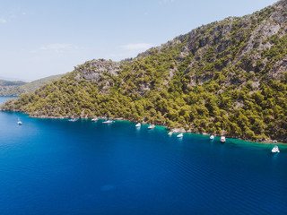 Wall Mural - Aerial drone perspective of luxury Turkish gulets and yachts in the deep blue and turquoise waters of the mediterranean sea. The bay and cliffs protect the boats from the strong winds.