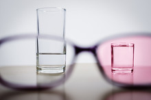 Optimism, Illustrated By Comparing Less Than Half Full Glass In One Frame Of Glasses With Full Smaller Glass Through Rose Tinted Lens In Another Frame. Amount Of Water Is About Same In Both Glasses.