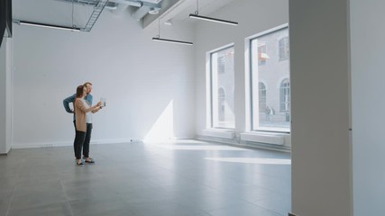 Wall Mural - Young Hipster Man and Female Stand in an Empty White Office and Map it with an Augmented Reality Software on a Tablet. Sunlight Shines Through Big Windows. Room Has Tracking Points for Video Software.