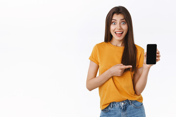 Wall Mural - Excited, thrilled young pretty woman in yellow t-shirt, showing smartphone screen, stare amazed and amused, smiling fascinated, pointing mobile display, found amazing app, white background