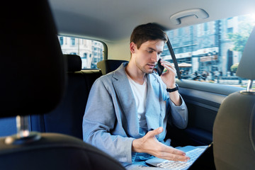 Wall Mural - Young brunette man in grey suit is riding in taxi in back seat of automobile. Businessman is talking on smartphone, working on laptop on way to meeting. Concept of fast rhythm of modern city.