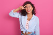Cheerful woman showing peace sign with her fingers, beautiful young female with curly hair showing victory sign and looks smiling directly at camera isolated over pink background. Gesture concept/