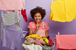 Funny curly housewife tells secret, smiles broadly, makes hush gesture, does washing at home, stands near pile of linen, hangs washed clothes on clothesline, isolated over purple background.