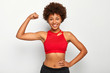 © WHstudio Leushin N - Horizontal shot of positive dark skinned woman shows biceps, demonstrates strong hand, has slim figure, wears sport bra, smiles pleasantly, isolated over white background. People and strength