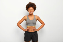 Photo Of Optimistic Dark Skinned Sporty Woman Keeps Hands On Waist, Smiles Happily, Dressed In Sport Bra And Black Shorts, Isolated Over White Background, Has Fitтess Training With Instructor