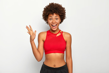 Photo of cheerful dark skinned woman raises palm, feels glad after workout, wears red sport bra, shows belly, has Afro hairstyle, leads healthy lifestyle, poses indoor over white background.