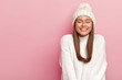 © wayhome.studio  - Horizontal shot of pretty young female with dark hair, keeps eyes shut, imagines pleasant moment, smiles pleasantly, shows white perfect teeth, enjoys comfort in new bought sweater, warm hat