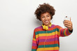 © WHstudio Leushin N - Cheerful dark skinned woman with dark curly hair, makes selfie via smartphone, dressed in colorful sweater, smiles gently, being music meloman, stands against white background, blank space aside
