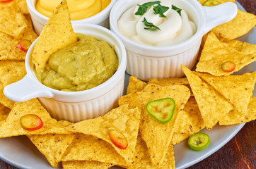 Nachos - yellow corn chips with various sauces in bowls: guacamole, cheese sauce, white sauce, on a wooden table. Mexican food concept.