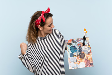 Wall Mural - Young artist woman holding a palette over isolated blue background celebrating a victory