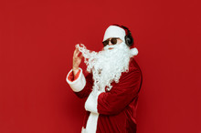 Studio Photo Of Fashionable Santa Claus In Sunglasses And Headphones Stands On A Red Background And Plays With A Beard With A Serious Face. Isolated Portrait Of Beautiful Santa. Christmas Concept.