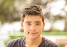 Close Up Portrait Of A Young  Teenager Man Looking At Camera With A Wondering Expression On His Face, Against A Green Blur Background. Outdoors Portarits.