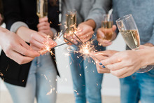 Hands Of Young Friends Holding Burning Bengal Lights And Flutes Of Champagne