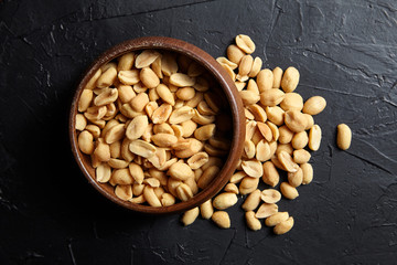 Round wooden bowl with roasted peeled peanuts, salty beer snack on dark background, top view