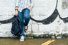 Front View Of Cool Looking And Stylish Teenager's Body Stand And Pose Against White Brick Wall With Black Spray Painted Graffiti Art In City, Wear Jean And Long Sleeve Shirt, Hold Backpack, Waiting.