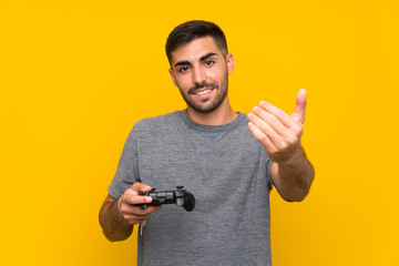 Wall Mural - Young handsome man playing with a video game controller over isolated yellow background inviting to come with hand. Happy that you came