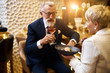 © alfa27 - Mature couple of caucasian man and woman sit in restaurant and eat dessert, drink glass of wine. Male in tuxedo, female in white blazer. Woman holding dish