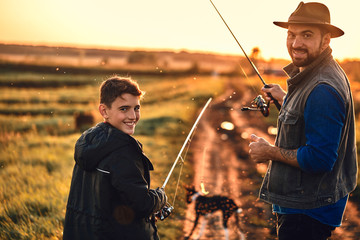 Father and son took fishing rods, fishing tackle and dog. They go on country road near lake for fishing. They smile and look at camera. Sunset.