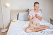 © alfa27 - Pretty short-haired girl woke up and drink tea. Female in t-shirt and shorts with crossed legs on bed. Background light room.