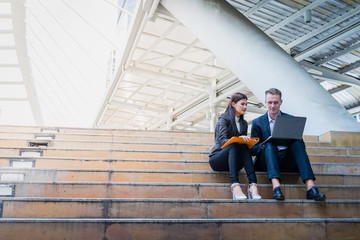 Asian business woman and Caucasian businessman sit on the staircase and hold a paper of financial report with laptop. concept of sale report and marketing plan with copy space for text.