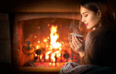 Beauty young Christmas woman sitting near fireplace in dark room at home and drinking hot beverage from a big cup. Beauty girl Enjoying tea, coffee or hot chocolate in a cozy room. Xmas scene