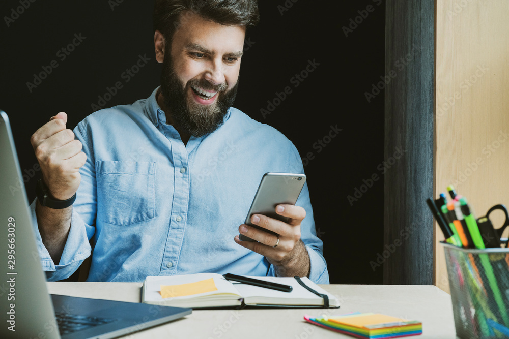 Man rejoices in victory while looking at screen of smartphone. Worker at desk with laptop and notebook. Male getting high-paid job - obrazy, fototapety, plakaty 