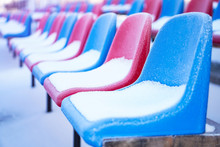 Snow-covered Multi-colored Seats In The Stadium In Winter. Snowfall Interferes With Sports. Uncleaned Stadium. Ecological Disaster Freeze