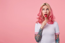 Portrait Of Attractive Young Tattooed Lady With Pink Curly Hair Looking Aside Dreamily, Posing Over Pink Background In Casual White T-shirt And Licking Candy On Stick