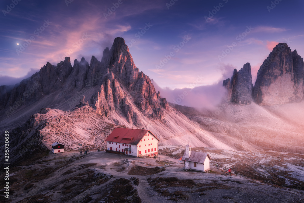 Mountains with beautiful house and church at sunset in autumn. Landscape with buildings, high rocks, purple sky with moon and clouds, sunlight. Mountains in fog. Tre Cime park in Dolomites, Italy - obrazy, fototapety, plakaty 