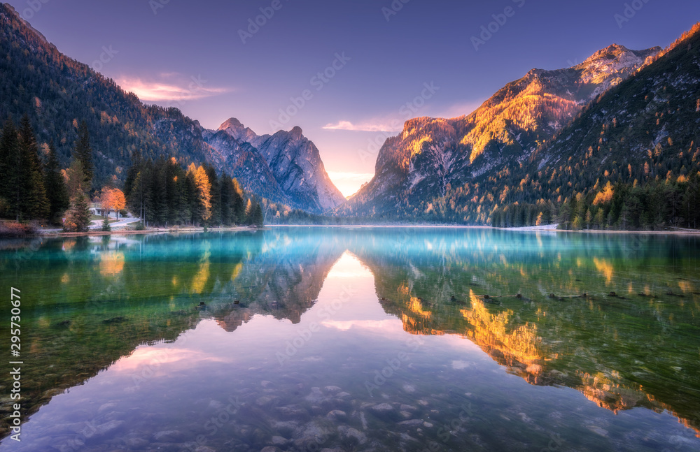 Mountain lake at sunrise in autumn. Landscape with lake, purple sky with gold sunlight, blue fog over the water, reflection, trees with colorful leaves, high rocks in fall. Forest in Dolomites, Italy - obrazy, fototapety, plakaty 