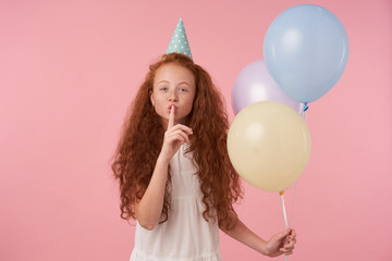 Wall Mural - Portrait of curly long haired little girl in festive clothes and birthday cap posing over pink background, keeping forefinger on her lips and asking to keep secret. Children and celebration concept