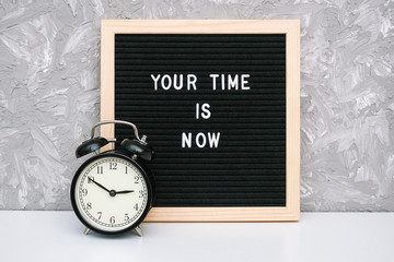 Wall Mural - Your time is now. Motivational quote on letter board and black alarm clock on table against stone wall. Concept inspirational quote of the day