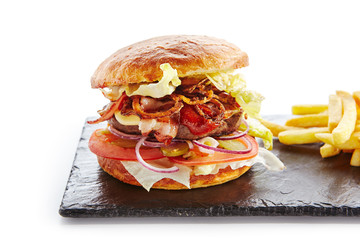 Canvas Print - Fresh Beef Burger with Bacon, Fried Onions, Tomato Sauce, Pickled Cucumbers, Green Lettuce, Cheese and French Fries Garnish. Hamburger or Beefburger on Natural Black Stone Plate Isolated on White