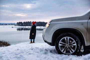 young adult woman in winter clothes with red hat with bubo standing near suv car at river side with beautiful view