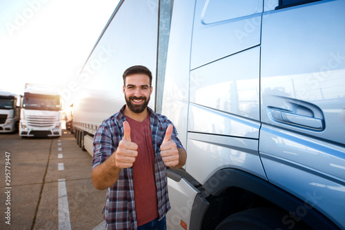 Middle aged professional trucker holding thumbs up and standing by his truck. Happy truck driver. Transportation services.