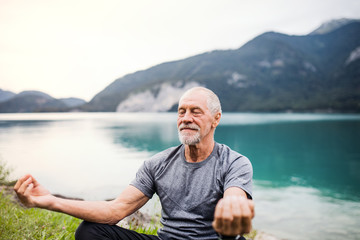 Sticker - A senior man pensioner sitting by lake in nature, doing yoga exercise.