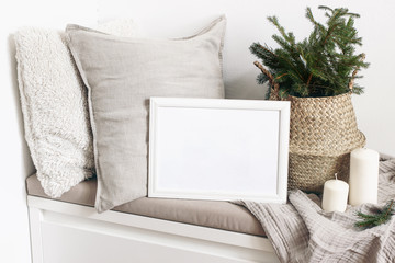 White blank wooden frame mockup with Christmas tree, candles, linen cushions and plaid on the white bench. Poster product design. Scandinavian home decor, nordic design. Winter festive concept.