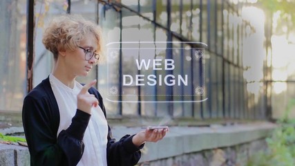 Curly young woman in glasses interacts with a hud hologram with text Web Design. Blonde girl in white and black clothes uses technology of the future mobile screen