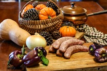 Selective Focus Still Life Of Preparation Of German Bratwurst Purple Peppers And Onions On Wooden Cutting Board With A Large Knife, With Blurred Basket Of Pumpkins And Pine Cones In Background