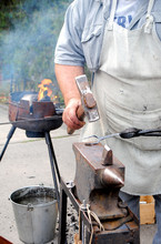 Blacksmith. Forging A Medieval Fork On An Anvil. A Blacksmith Forges Outdoors Next To A Brazier To Heat The Metal.