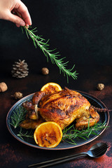 Roasted whole chicken for Christmas or New Year on a dark background with Christmas decorations. Baked Chicken with Oranges and rosemary. Christmas dinner. Female hand decorating chicken with rosemary