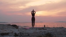The Camera Is Going Towards A Young Woman Doing Namaste Pose Before Starting Her Yoga Routine. The Beautiful Seaside With Pink Sunset Colors Surrounds Her.