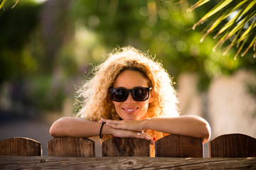 Wall Mural - Portrait of beautiful caucasian young blonde curly woman cheerful looking at the camera with sun lights in background and defocused green natural outdoor background - happy middle age lady concept