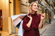 © Anton - Cheerful blond girl in knitted sweater with shopping bags on shoulder happily looking away on street