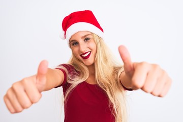 Young beautiful woman wearing Christmas Santa hat over isolated white background approving doing positive gesture with hand, thumbs up smiling and happy for success. Winner gesture.