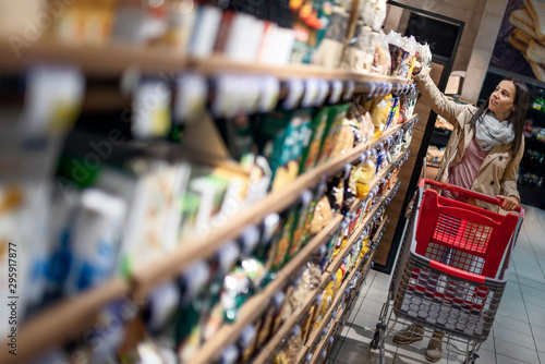 Fotomural Gorgeous young woman with a shopping cart looking at some products on a supermarket aisle
