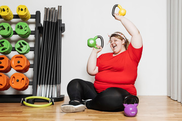 Wall Mural - Fat and funny girl dressed in the sports wear is working out with dumbbells in the gym next to other sports equipment