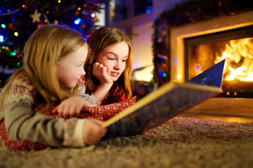 Wall Mural - Happy young sisters reading a story book together by a fireplace in a cozy dark living room on Christmas eve. Celebrating Xmas at home.