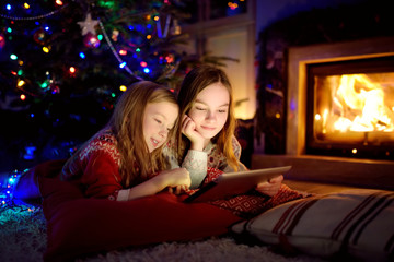 Wall Mural - Two cute young sisters using a tablet pc at home by a fireplace in warm and cozy living room on Christmas eve.