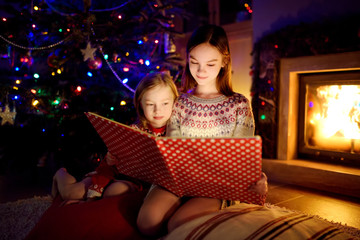 Wall Mural - Happy young sisters reading a story book together by a fireplace in a cozy dark living room on Christmas eve. Celebrating Xmas at home.